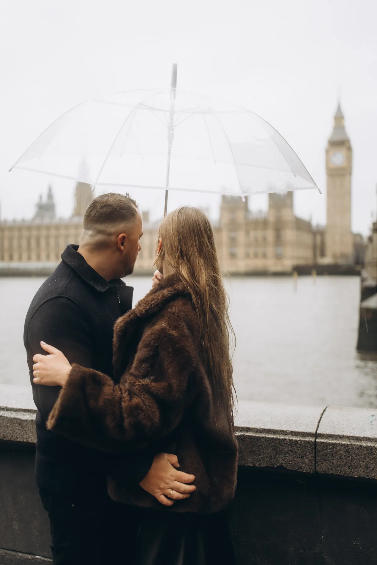 Couple seen from behind under a clear umbrella, looking across the Thames at Big Ben