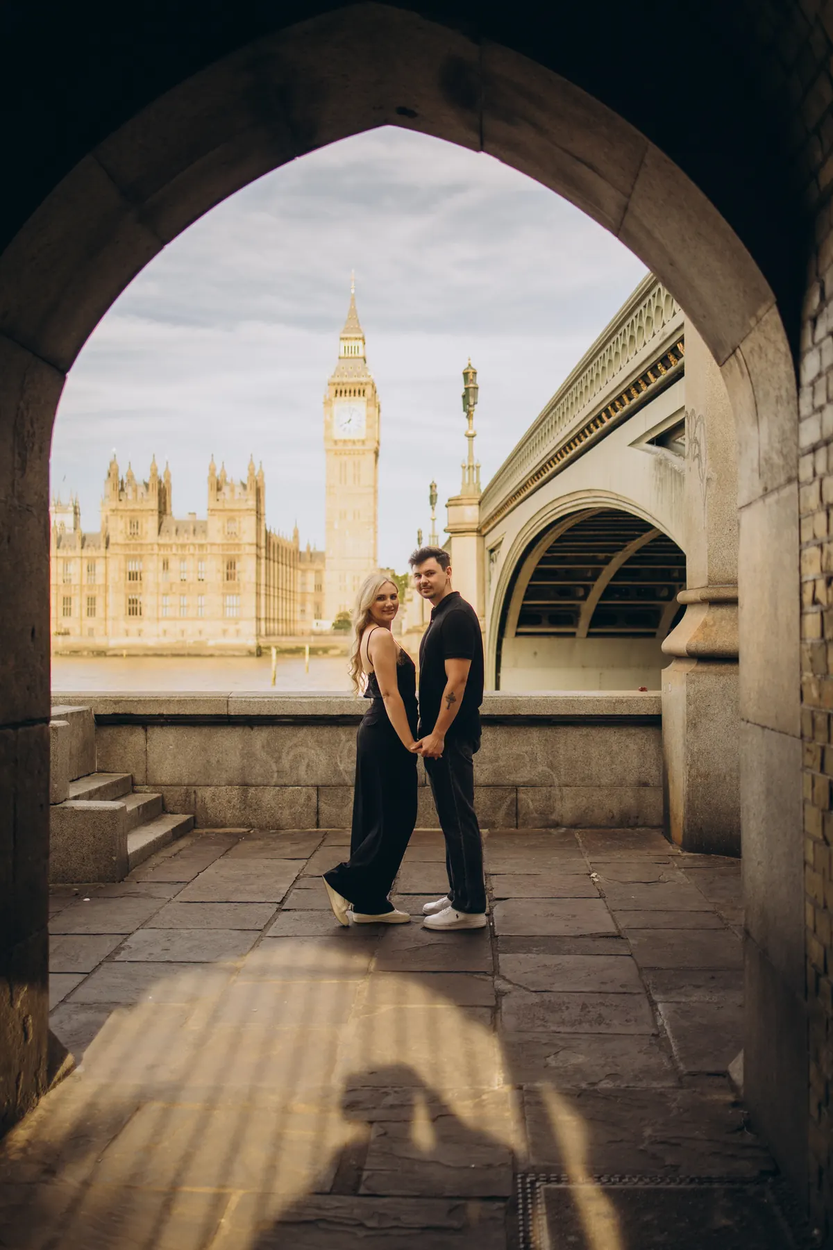 Couple holding hands inside the arch under Westminster Bridge with Big Ben across the river