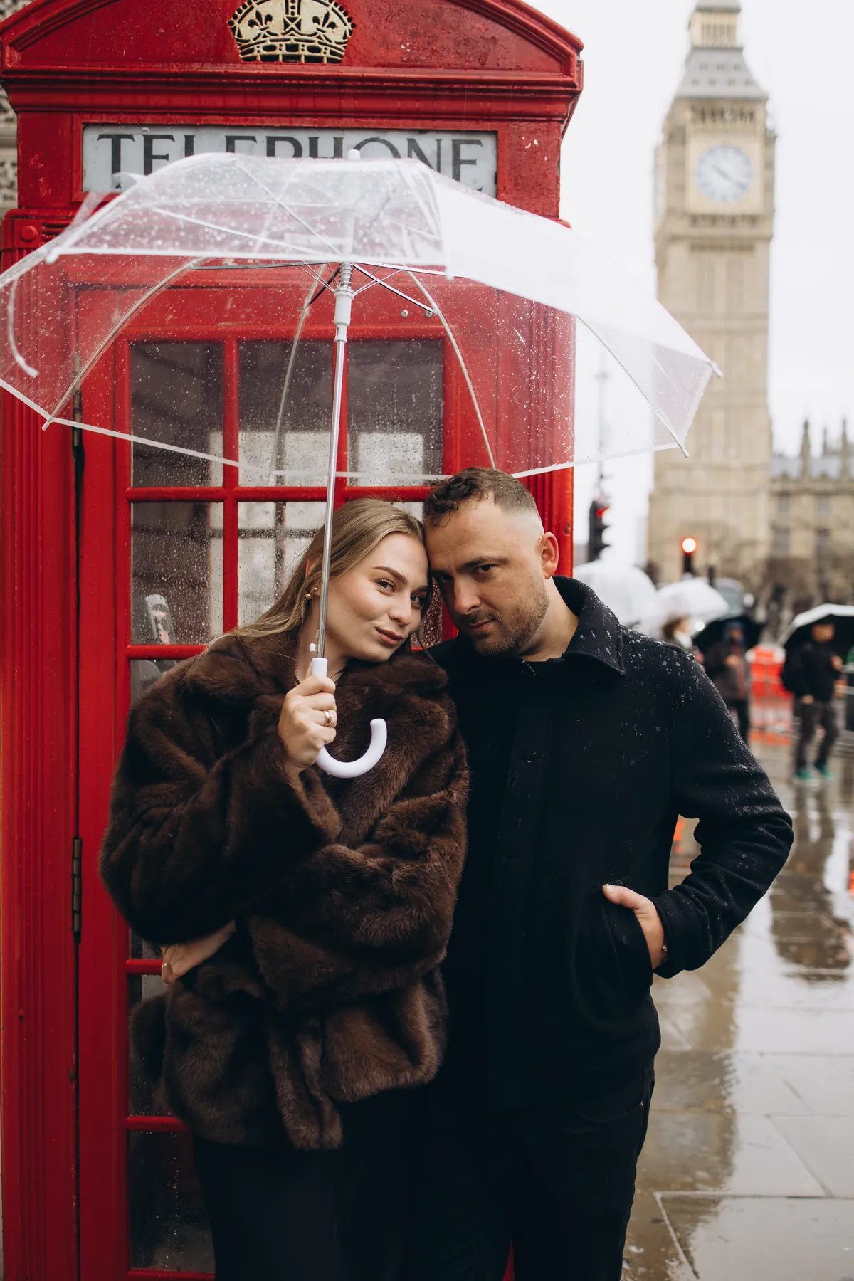 Couple leaning heads together under a clear umbrella in front of a red phone box and Big Ben