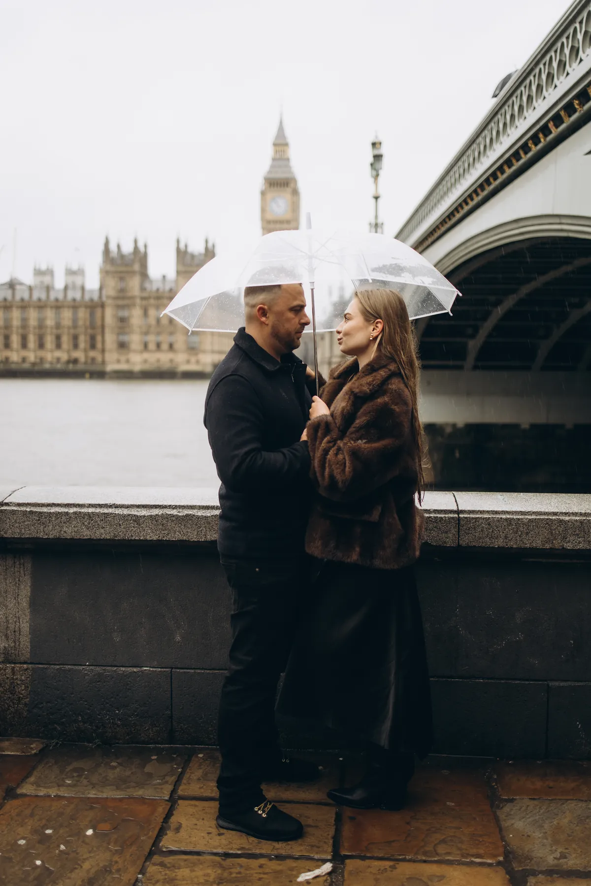 Couple facing each other under a clear umbrella on the South Bank with Big Ben across the Thames