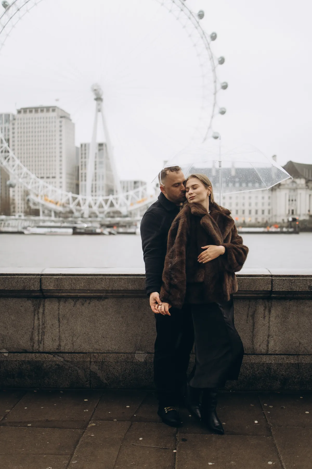 Man holding his partner from behind under a clear umbrella by the Thames, the London Eye across the river