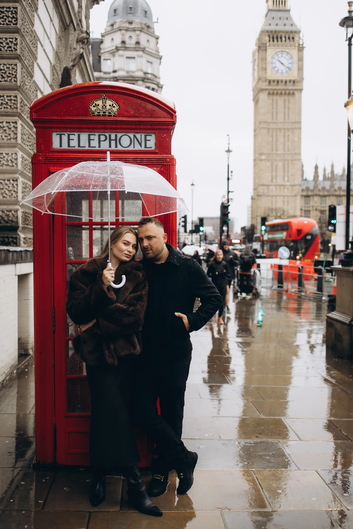 Full-length photo of a couple by a red phone box on a wet pavement with Big Ben and a red bus behind