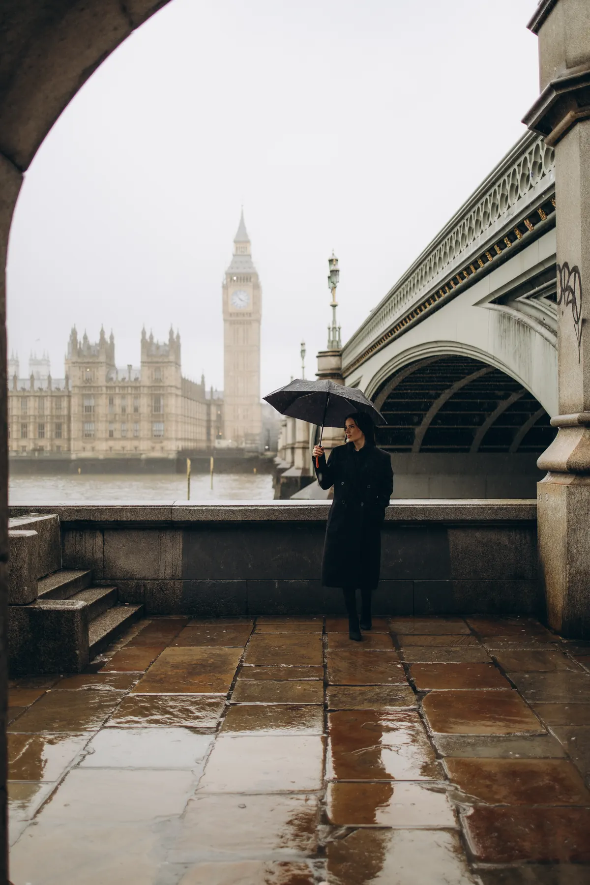 Woman with an umbrella on the wet South Bank framed by an arch, Big Ben behind
