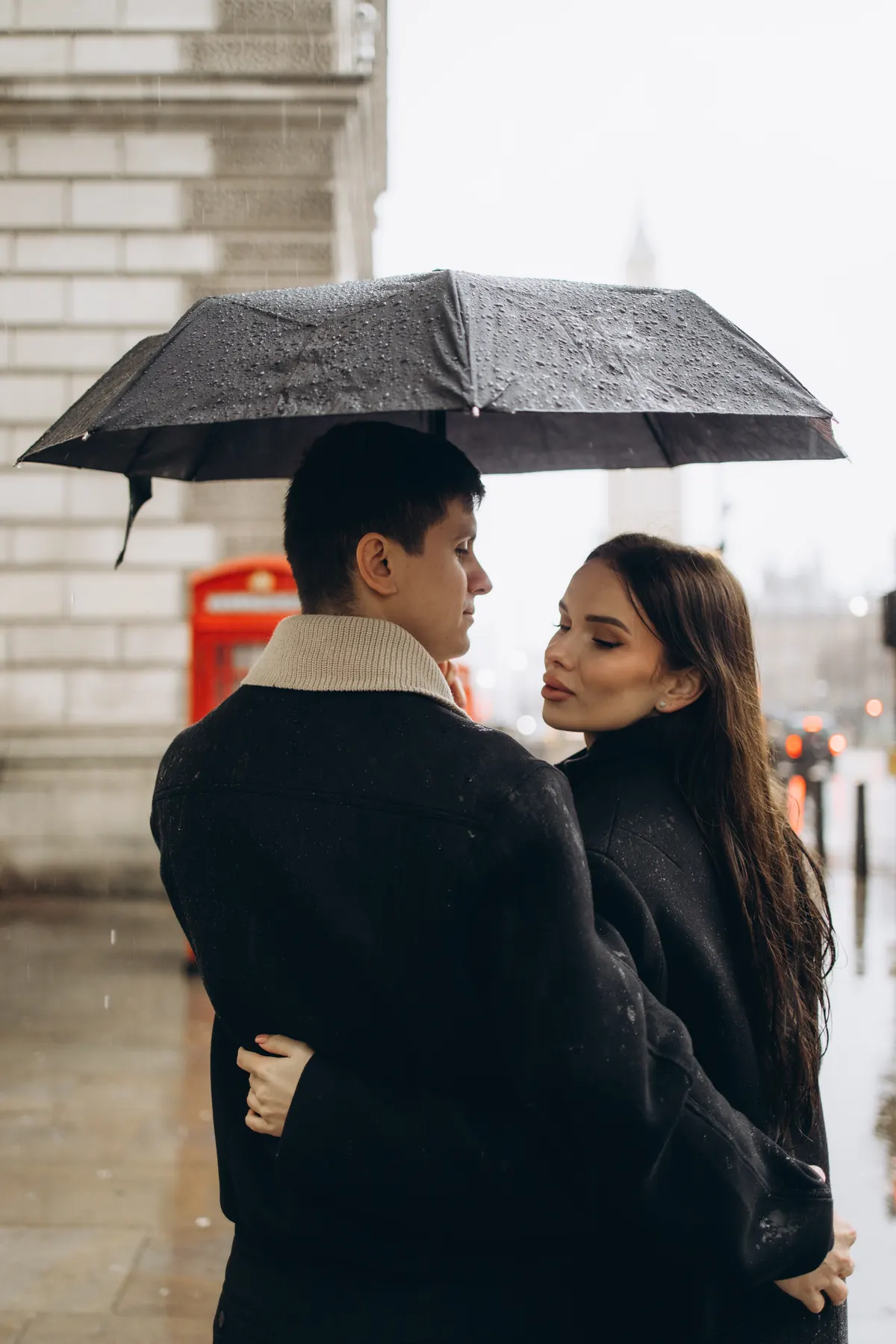 Couple under a black umbrella in the rain near a red phone box and Big Ben