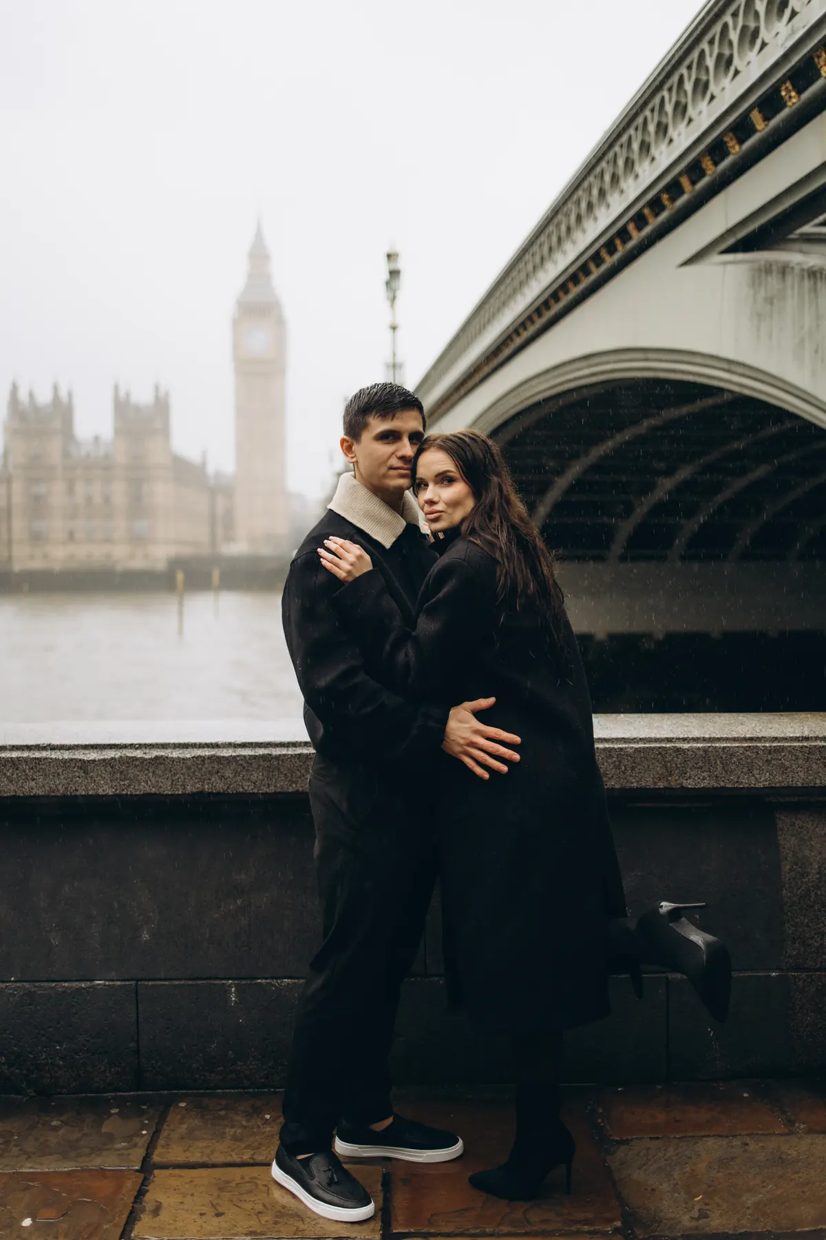 Couple embracing by the river wall under Westminster Bridge in the rain, Big Ben in the mist
