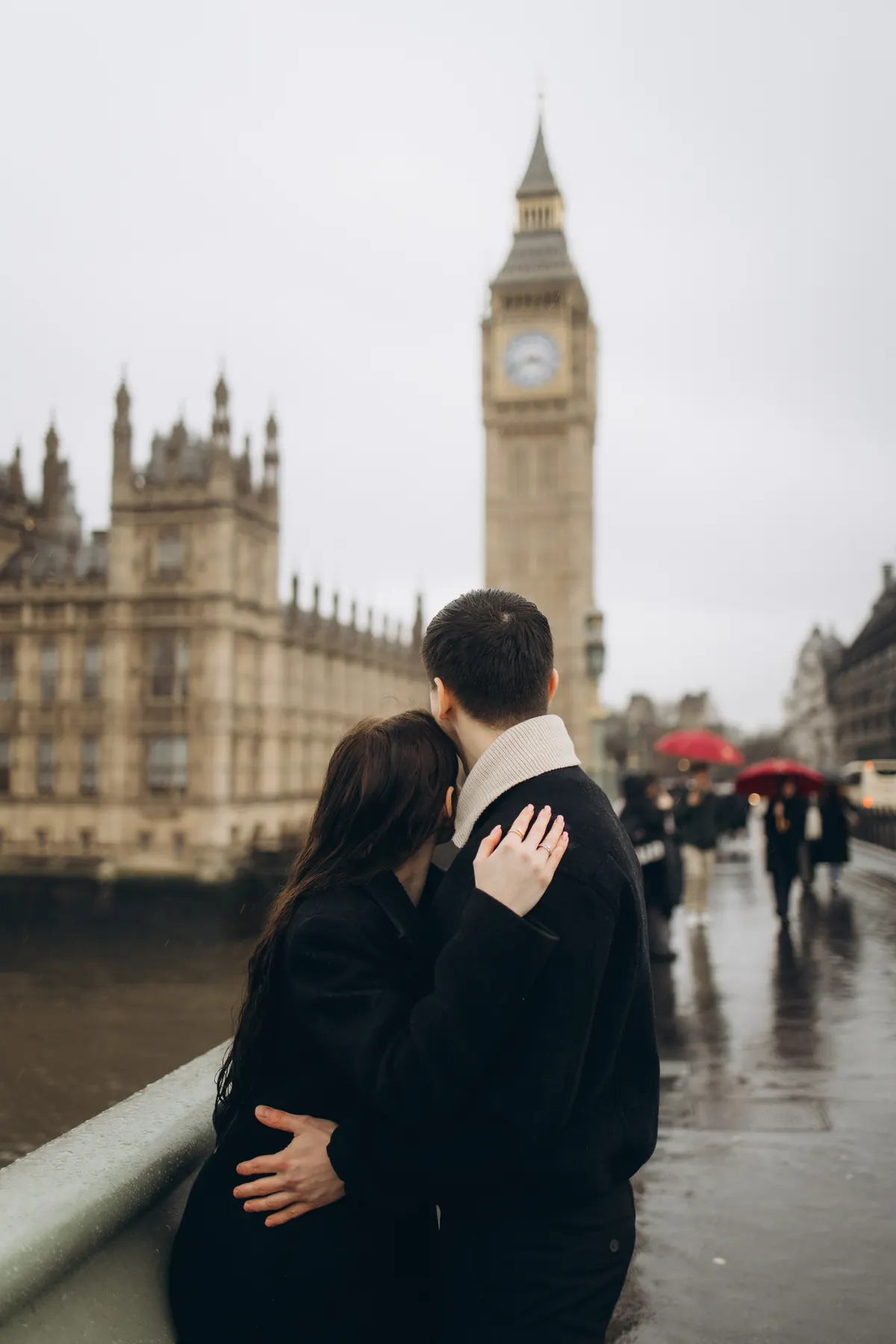 Couple hugging on wet Westminster Bridge looking at Big Ben, red umbrellas behind