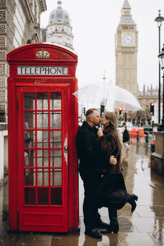 Couple kissing under a clear umbrella by a red phone box with Big Ben behind them in the rain
