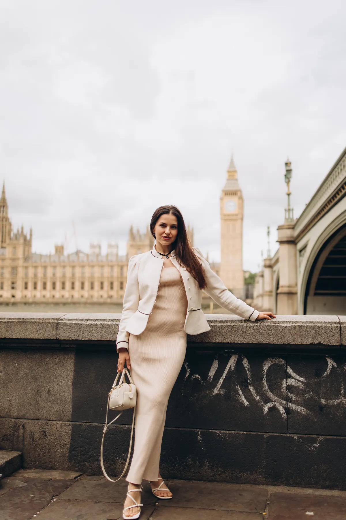 Woman in a cream jacket by the South Bank river wall facing Big Ben