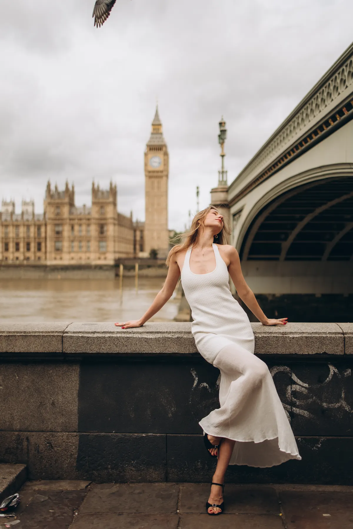 Woman in a white dress on the river wall as a pigeon flies past Big Ben