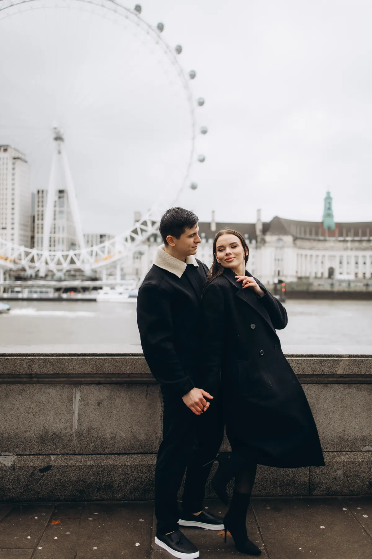 Couple in black coats by the Thames with the London Eye behind on a grey day