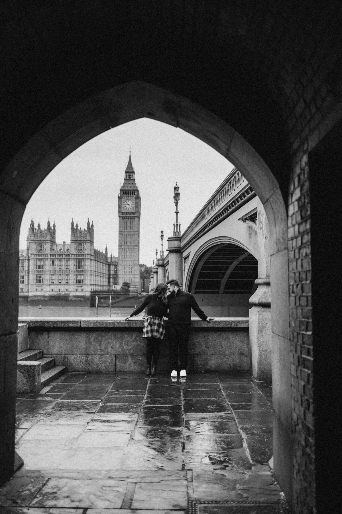 Black and white photo of a couple framed by the arch under Westminster Bridge with Big Ben across the river