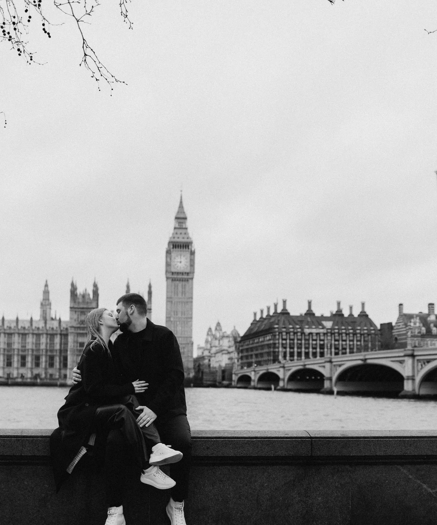 Black and white photo of a couple kissing on the river wall opposite Big Ben