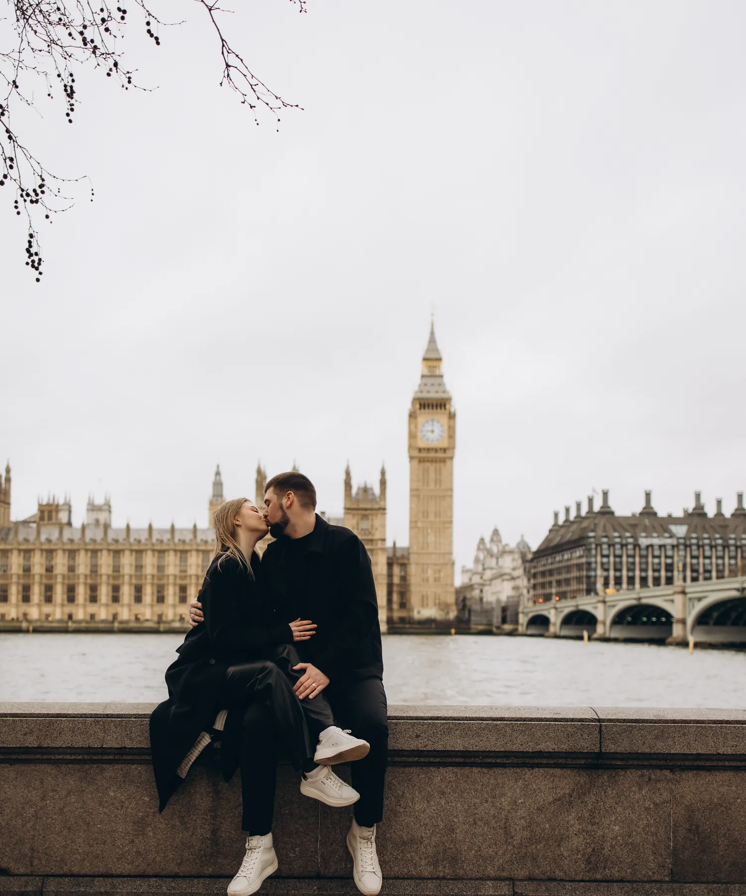 Couple kissing while sitting on the river wall with Big Ben, the Houses of Parliament and Westminster Bridge across the Thames