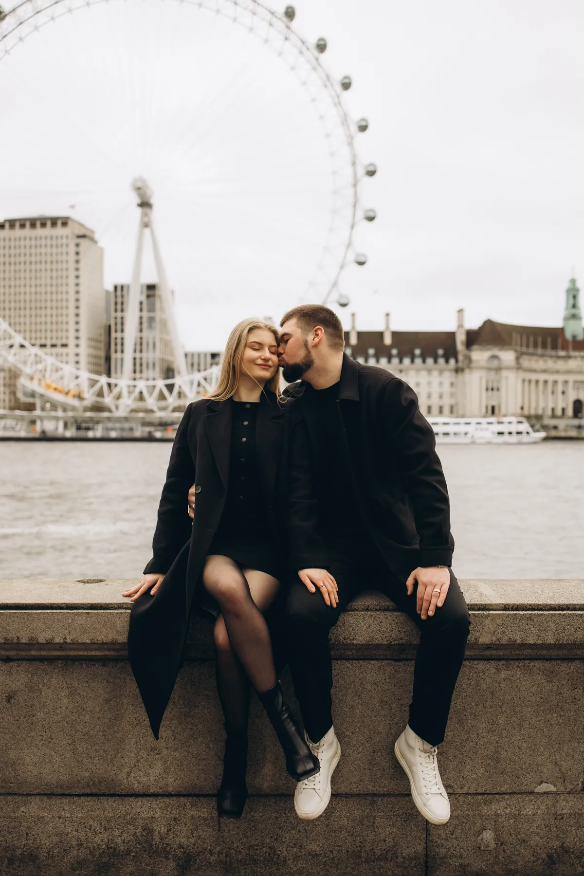 Man kissing his partner on the cheek as they sit on the river wall opposite the London Eye