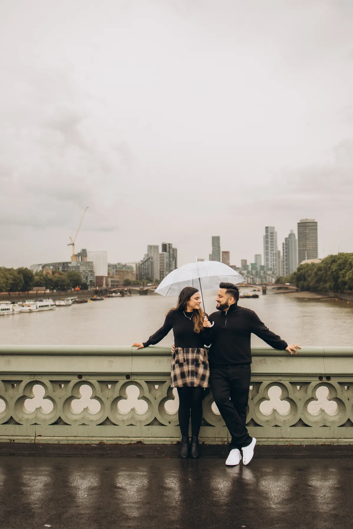 Couple under a clear umbrella leaning on the Westminster Bridge railing, the Thames behind them
