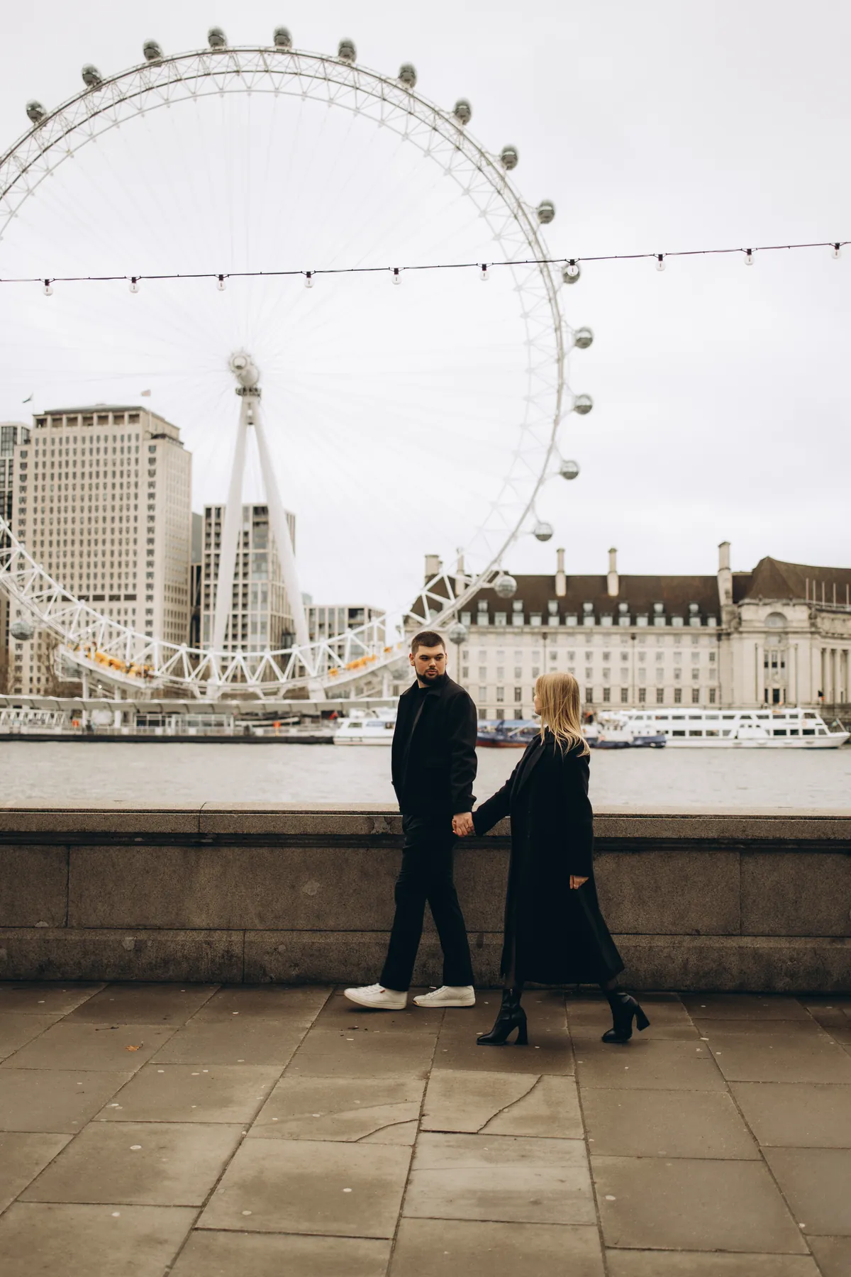 Couple in black coats walking hand in hand along the river wall with the London Eye across the Thames