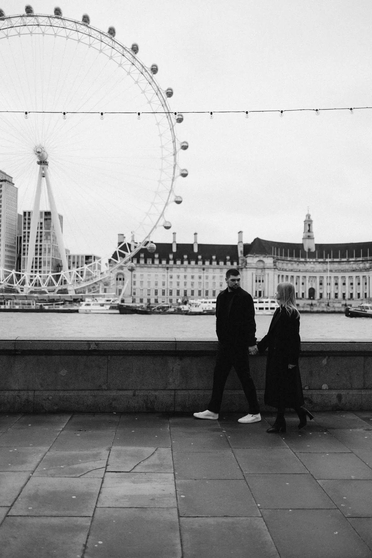 Black and white photo of a couple walking hand in hand by the Thames with the London Eye behind