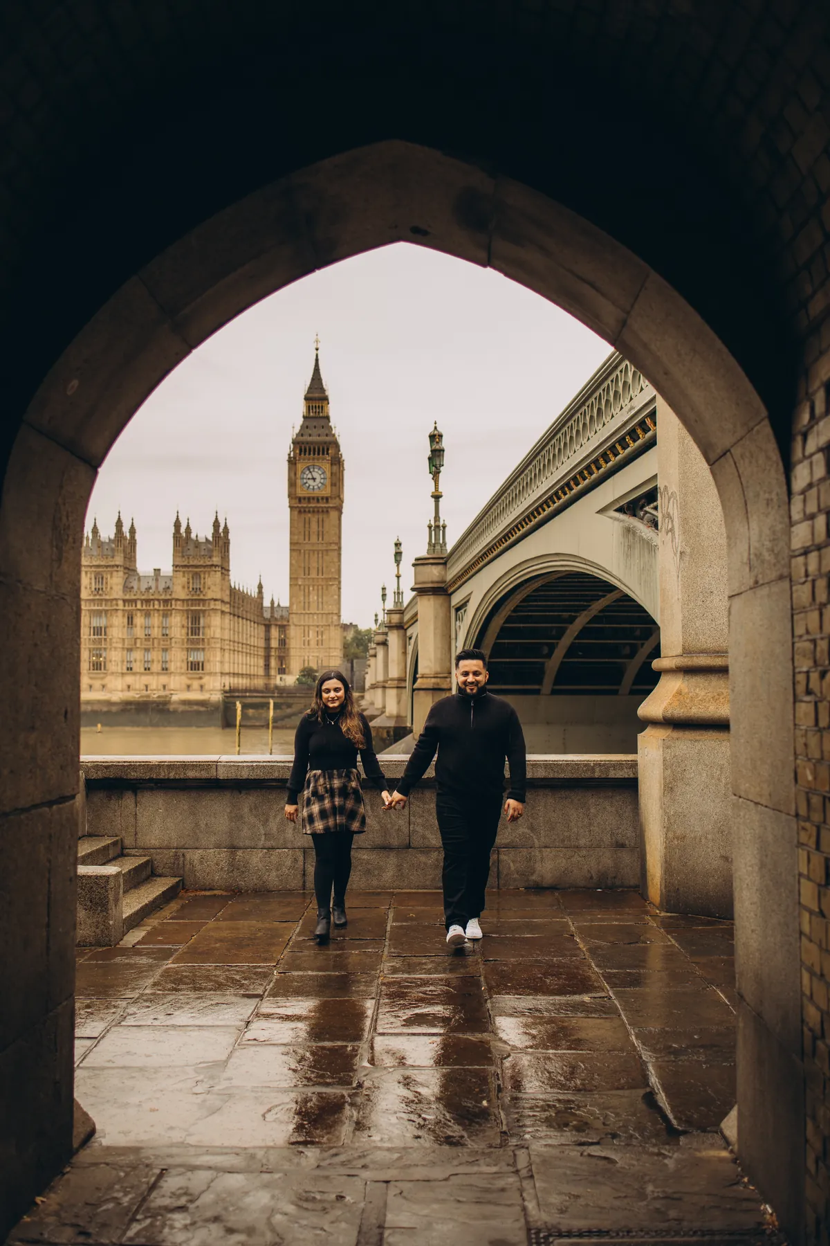Couple walking hand in hand out of the arch under Westminster Bridge with Big Ben across the river