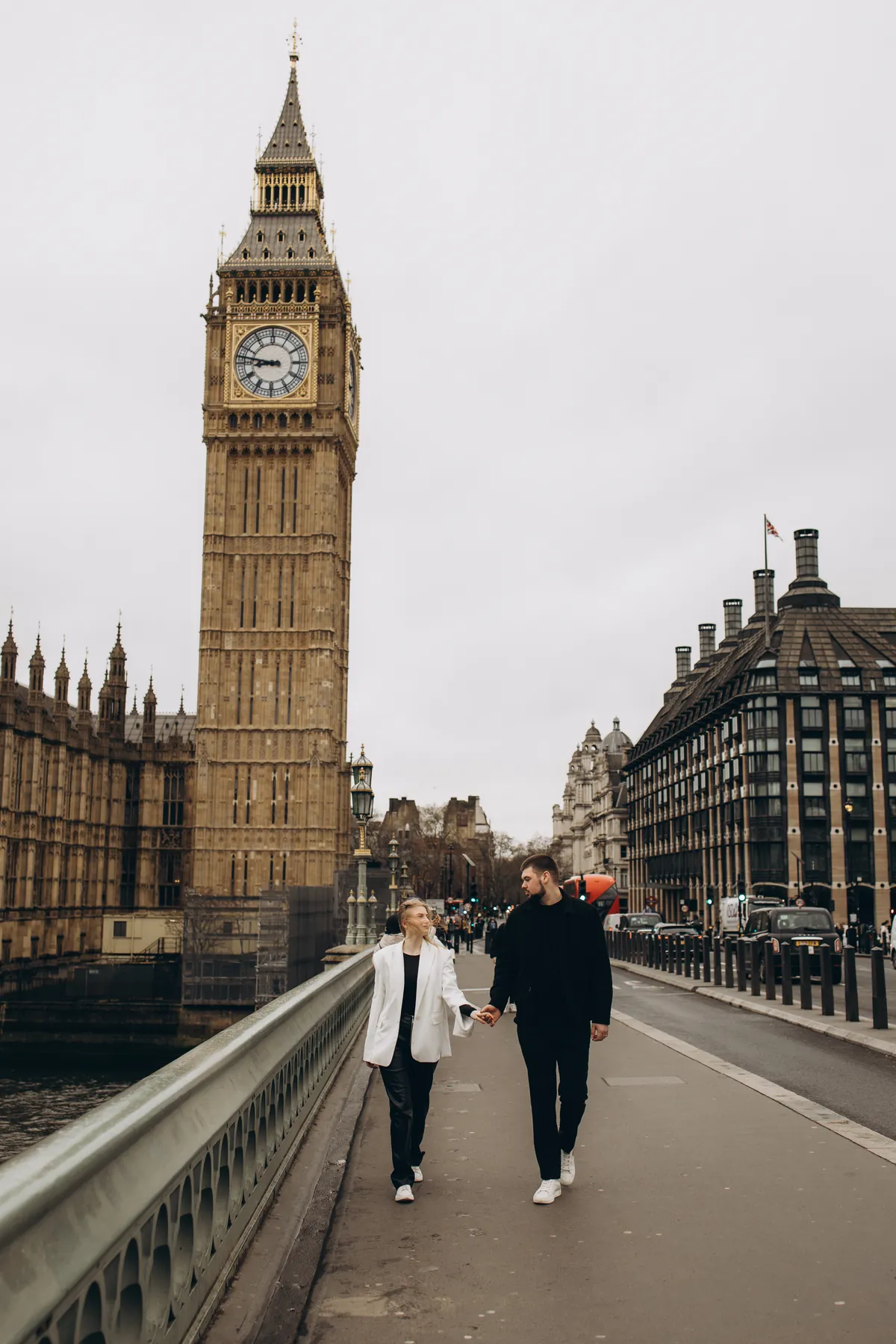 Couple walking hand in hand on Westminster Bridge with Big Ben behind, she in a white blazer