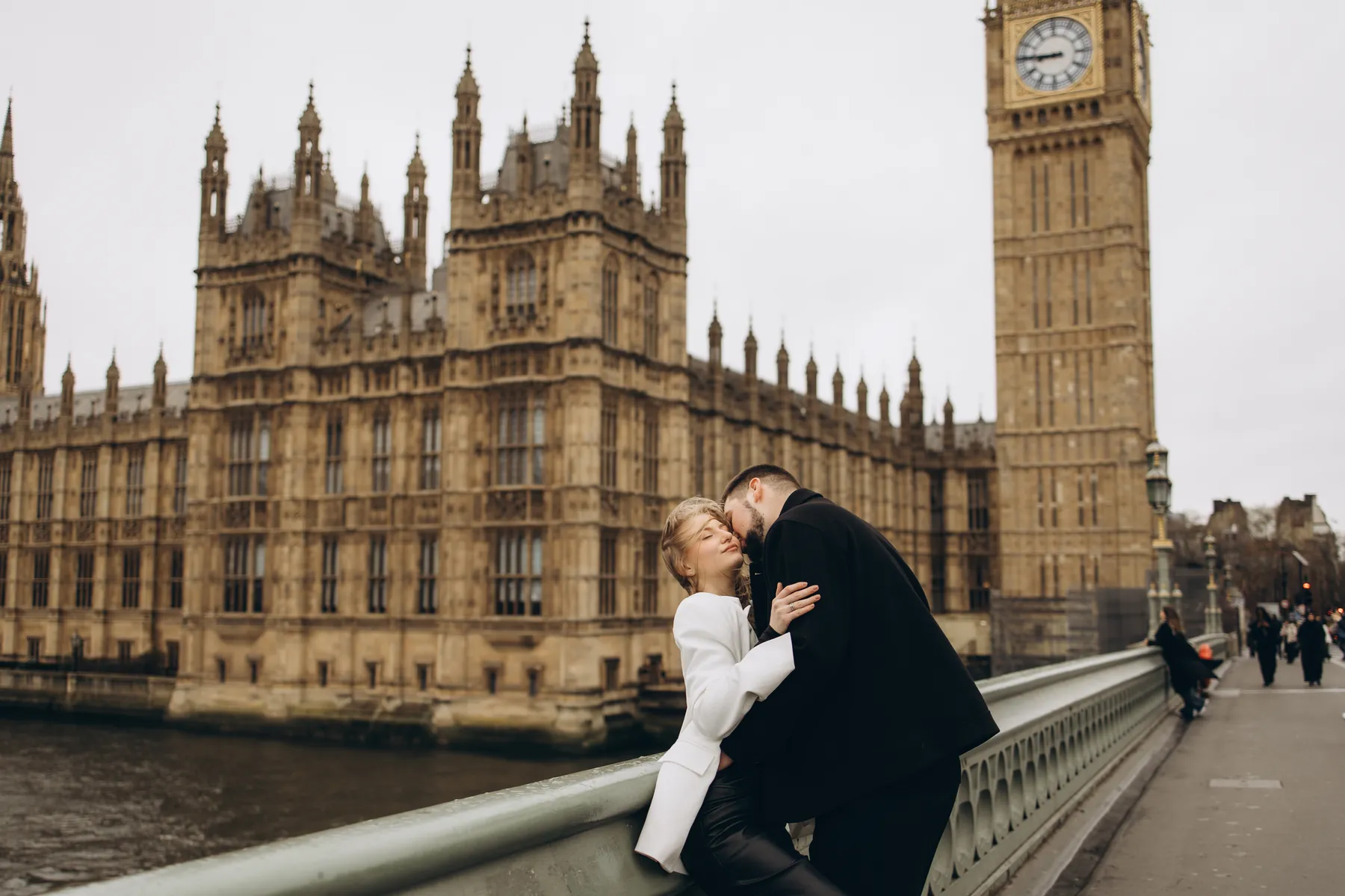 Couple embracing on Westminster Bridge, she in a white blazer, with the Houses of Parliament and Big Ben behind