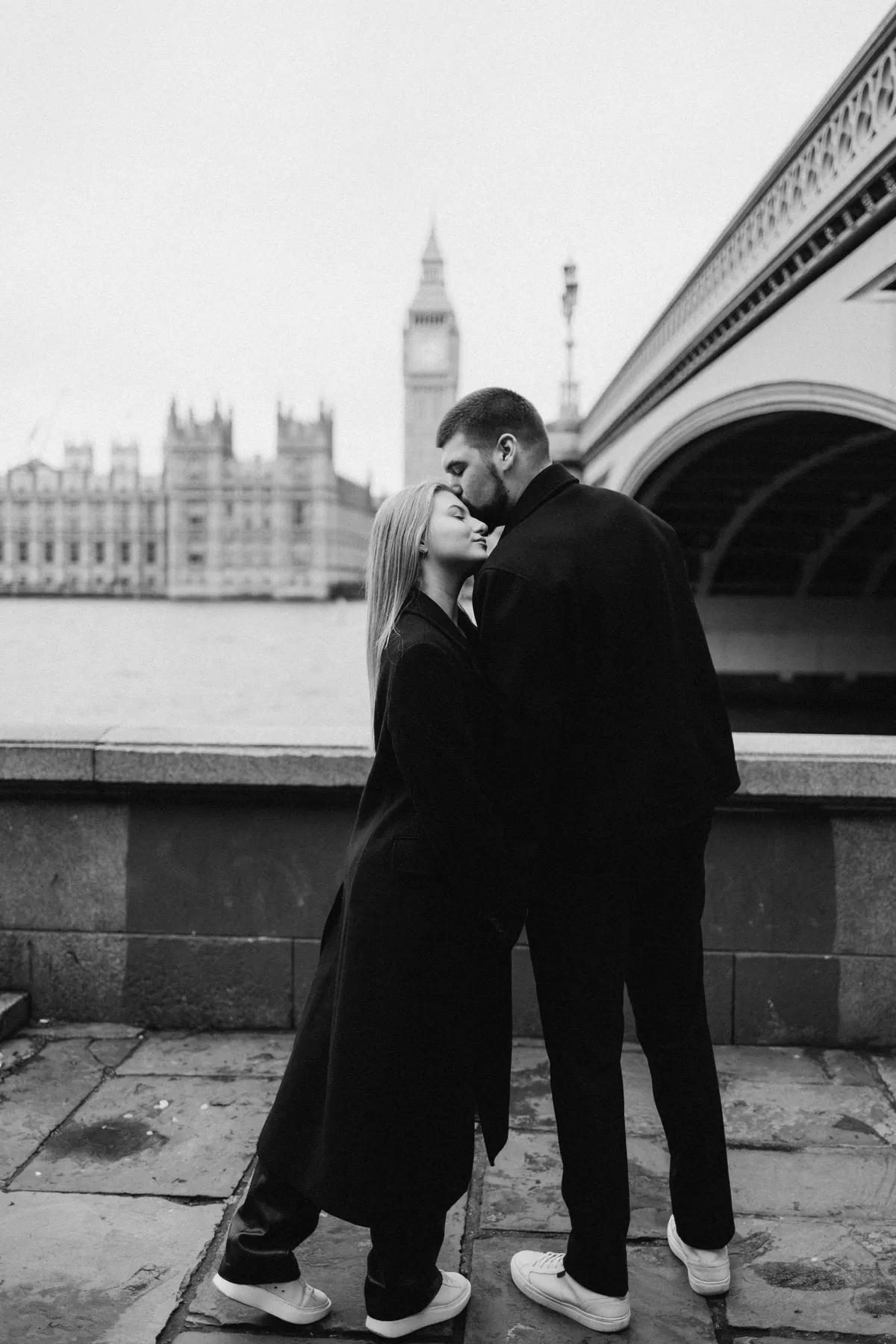 Black and white photo of a man kissing his partner on the forehead by the river wall with Big Ben behind
