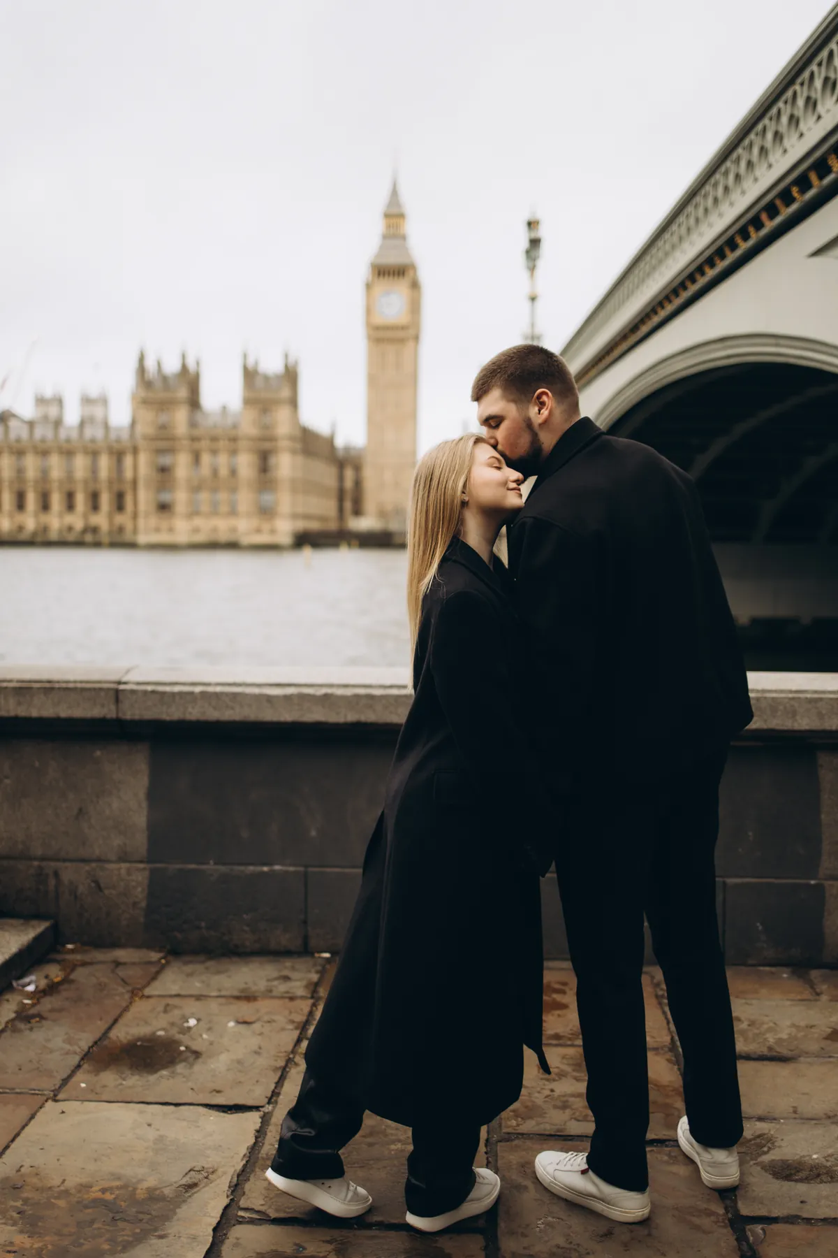 Man kissing his partner on the forehead on the South Bank with Big Ben and Westminster Bridge behind