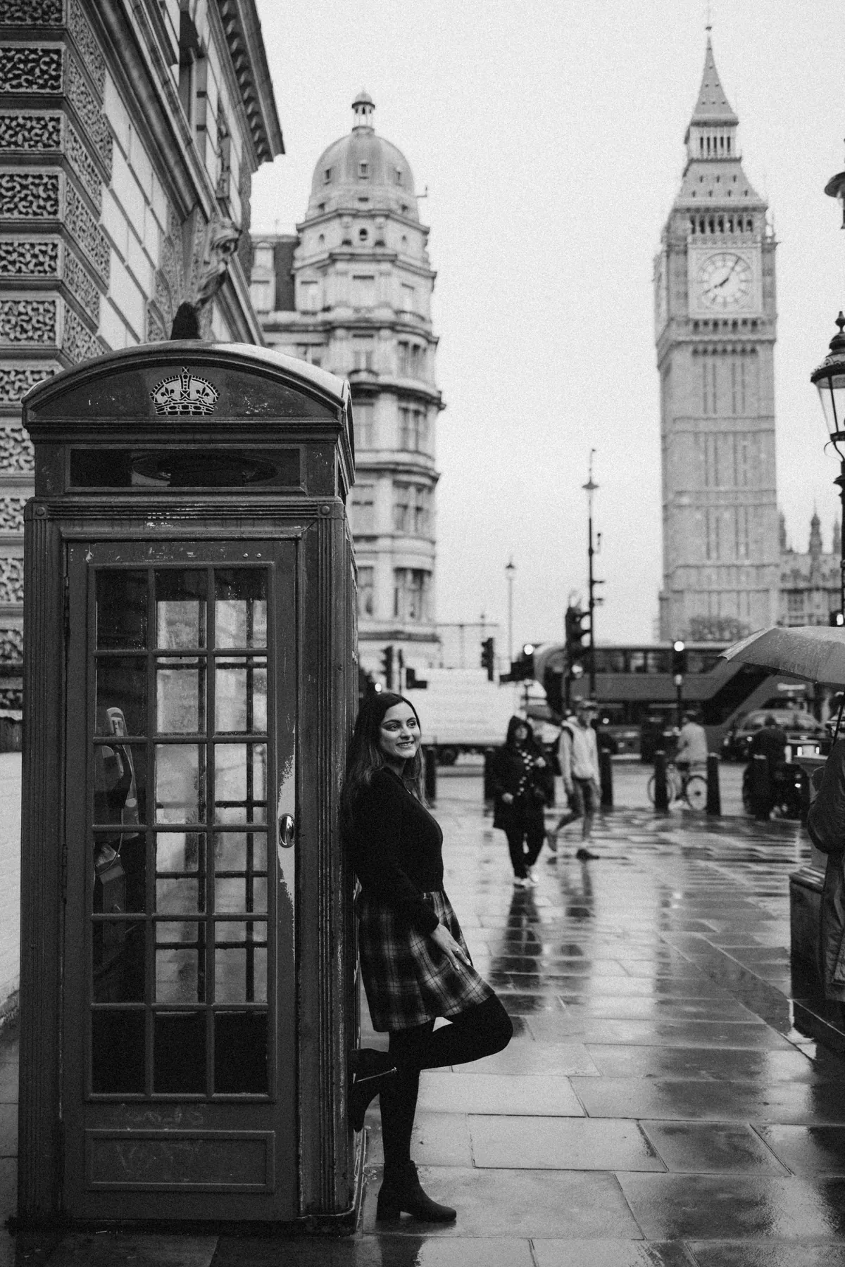 Black and white portrait of a woman leaning on a phone box with Big Ben on a rainy street
