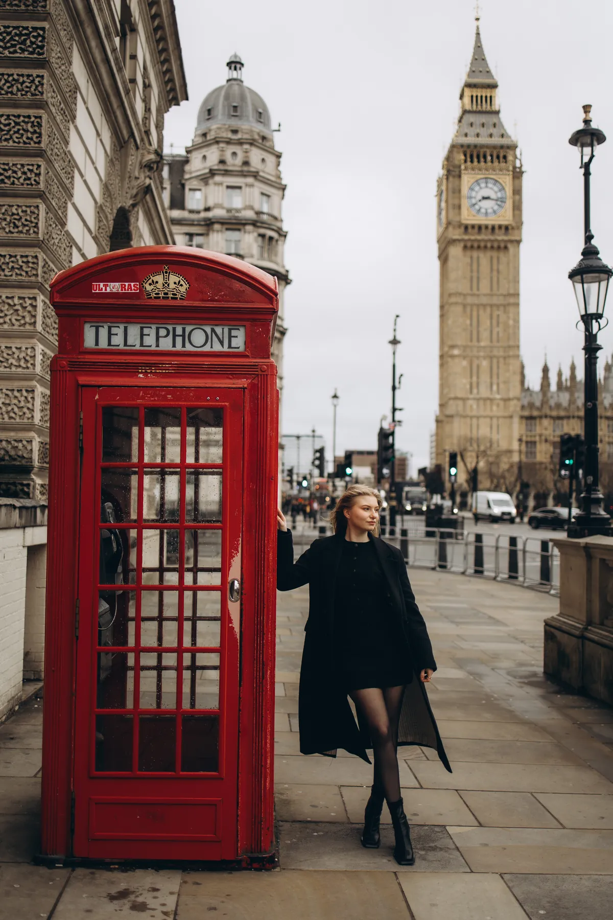 Woman in a long black coat leaning on a red phone box with Big Ben behind